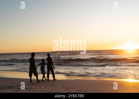 Silhouette ragazzo afroamericano con madre e padre che camminano sulla spiaggia contro il cielo al tramonto Foto Stock