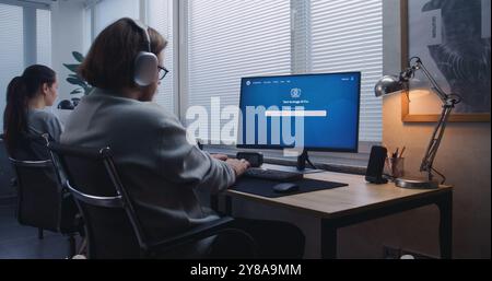 Uomo adulto con cuffie di tipo prompt in chatbot di intelligenza artificiale su PC. Il generatore di immagini AI crea un ritratto di alta qualità del robot fantascientifico. Una collega donna lavora al computer in background. Foto Stock