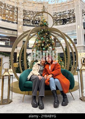 Madre e figlia si siedono comodamente su una panchina elegante di fronte all'albero di Natale decorato in un centro commerciale. l'ambiente festivo e i loro abiti caldi Foto Stock