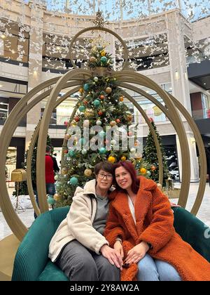 Madre e figlia si siedono comodamente su una panchina elegante di fronte all'albero di Natale decorato in un centro commerciale. l'ambiente festivo e i loro abiti caldi Foto Stock