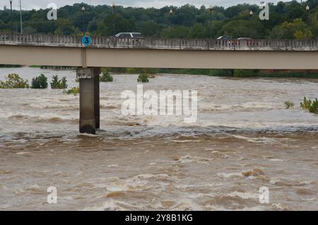Ampia visuale dall'alto verso il basso per un rapido movimento, slancinante e fangoso flusso d'acqua di fiume marrone argilla che rotola con tappi bianchi. Alberi verdi dal ponte Foto Stock