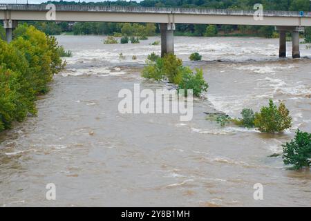 Ampia visuale dall'alto verso il basso per un rapido movimento, slancinante e fangoso flusso d'acqua di fiume marrone argilla che rotola con tappi bianchi. Alberi verdi dal ponte Foto Stock