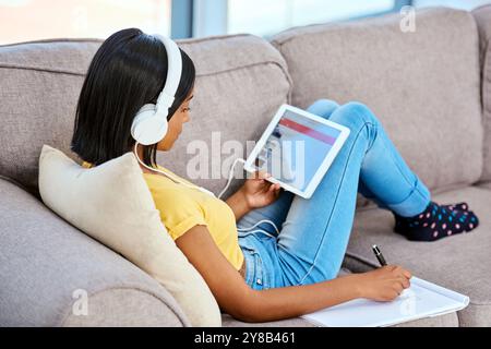 Ragazza, casa e tablet con cuffie e scrittura su notebook per lo studio, la revisione e la ricerca. Donna, adolescente e gen z sul divano in vita Foto Stock