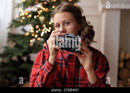 Una ragazza con un sorprendente vestito rosso, che tiene in mano una macchina fotografica d'epoca. Si siede di fronte a un albero di Natale splendidamente decorato, adornato da ghirlande scintillanti e luci scintillanti Foto Stock