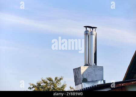 Due camini in stile moderno con forno e caminetto impilati in acciaio inossidabile sul tetto di una casa rurale ricoperti di ardesia sotto il cielo azzurro con spazio imbottito. Foto Stock