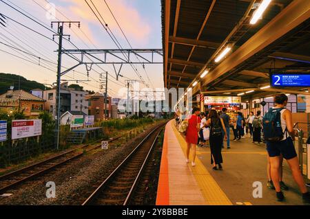 Una vivace scena in una stazione ferroviaria tra Yokohama e Tokyo. con pendolari in attesa sulla piattaforma mentre il sole tramonta. Foto Stock