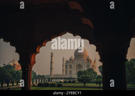 Il Taj Mahal è incorniciato da un arco ornato, con il suo marmo bianco che brilla di luce soffusa, circondato da giardini. Foto Stock