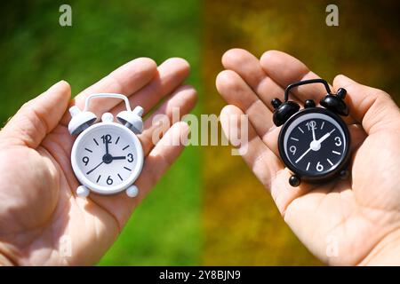 MONTAGGI FOTOGRAFICI, sveglie bianche e nere sono tenute con le mani, foto simboliche del cambio di orario in inverno, FOTOMONTAGE, Weißer und schwarzer Wecker W. Foto Stock