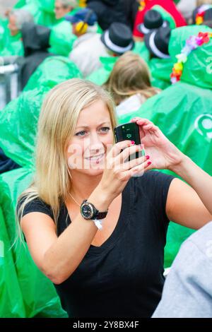 PRIMO SMARTPHONE, TEMPESTA ESTIVA, BEACH VOLLEY, MARIEHAMN, 2011: Una donna bionda scatta una foto con uno smartphone. La folla si copre con poncho antipioggia usa e getta mentre un'enorme tempesta estiva attraversa lo stadio con un diluvio di pioggia durante le semifinali dell'agosto 2011 al PAF Open di Mariehamn, Åland, Finlandia. Fotografia: Rob Watkins. INFO: Tra il 2009-2013 il PAF Open Beach Volley è stato un torneo annuale che si è tenuto a Mariehamn, Åland, Finlandia. Ha attirato le migliori squadre e giocatori internazionali come parte del FIVB World Tour ufficiale, mostrando alto livello Foto Stock