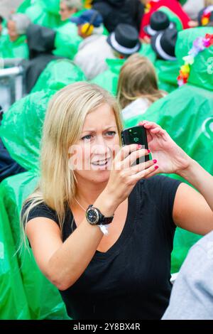 PRIMO SMARTPHONE, TEMPESTA ESTIVA, BEACH VOLLEY, MARIEHAMN, 2011: Una donna bionda scatta una foto con uno smartphone. La folla si copre con poncho antipioggia usa e getta mentre un'enorme tempesta estiva attraversa lo stadio con un diluvio di pioggia durante le semifinali dell'agosto 2011 al PAF Open di Mariehamn, Åland, Finlandia. Fotografia: Rob Watkins. INFO: Tra il 2009-2013 il PAF Open Beach Volley è stato un torneo annuale che si è tenuto a Mariehamn, Åland, Finlandia. Ha attirato le migliori squadre e giocatori internazionali come parte del FIVB World Tour ufficiale, mostrando alto livello Foto Stock