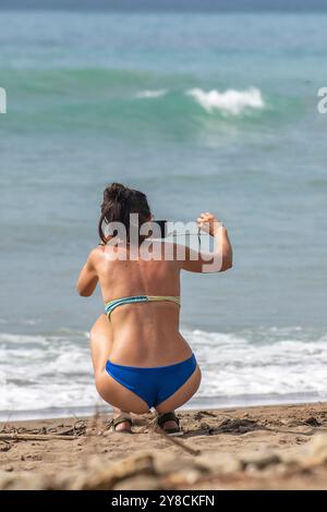 giovane donna attraente che indossa un bikini blu accovacciato sul bordo di una spiaggia mediterranea per scattare una foto delle onde che si infrangono Foto Stock