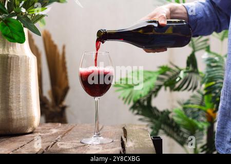 Un uomo irriconoscibile sta versando vino rosso francese versato in un bicchiere di bollicine con uno spruzzo sul tavolo di legno nel Garden Summer, primo piano Foto Stock