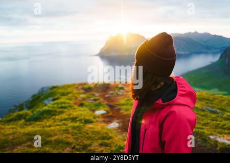 Una giovane donna con berretto marrone e giacca rossa osserva l'alba sulle montagne. Foto Stock