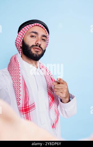 Uomo musulmano sicuro che indossa il tradizionale velo che punta con il dito e guarda la fotocamera anteriore dello smartphone. Un arabo serio parla in video mentre tiene in mano il cellulare fpv Foto Stock