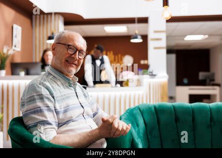 Ritratto di un anziano seduto su un comodo divano nell'esclusiva reception dell'hotel, in attesa della procedura di check-in. La confortevole lobby offre una rilassante area salotto per i viaggiatori di uomini anziani che guardano la macchina fotografica. Foto Stock