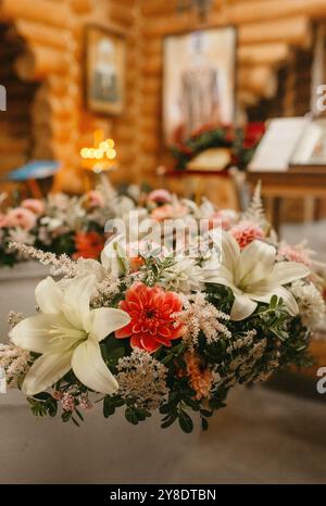 Un pezzo centrale di gigli bianchi, rose rosa e dahlias di corallo si trova in cima a un tavolo di legno all'interno di una chiesa, circondato da uno sfondo sfocato di gla macchiato Foto Stock