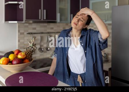 Donna che sente mal di testa e soffre di mal di testa in cucina a casa Foto Stock