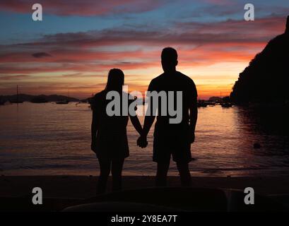 La coppia sagomata si trova vicino alla spiaggia, godendosi un momento romantico mentre il sole tramonta. Foto Stock