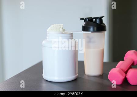 Primo piano di un frullato proteico in una bottiglia sul tavolo con manichini rosa e polvere di proteine del siero di latte bianco in una paletta. Sostituzione dei pasti dietetici per la perdita di peso. Foto Stock