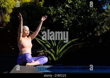 Donna birazziale felice e inalterata che pratica yoga seduto a bordo piscina in giardino, spazio fotocopie Foto Stock