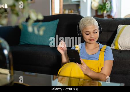 Donna birazziale alla moda e inalterata seduta a terra a casa con le cuffie che utilizzano lo smartphone Foto Stock