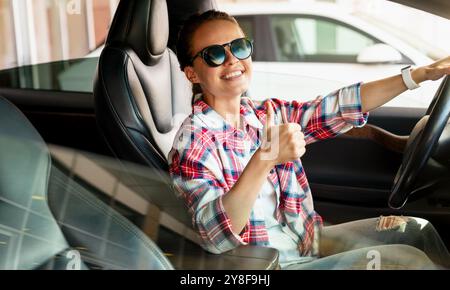 Una giovane donna siede nella sua nuova auto presso la concessionaria, sorridendo e dando un cartello con il pollice. Servizio di noleggio auto, car sharing. Foto Stock