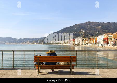 Vista posteriore di una donna e del suo cane che riposano su una panchina del Molo Mario Bestoso in una giornata di sole primaverili, Alassio (Savona), Liguria, Italia Foto Stock