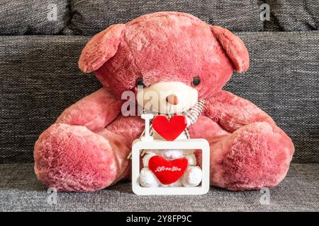Un grande orsacchiotto di peluche occupa il centro della scena, irradiando una tenue tonalità rosa. Di fronte all'orso, c'è una piccola scatola bianca con un coperchio trasparente. All'interno, a r Foto Stock