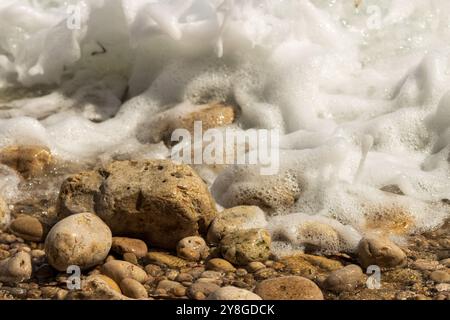 Questa immagine cattura una vista ravvicinata di una costa con schiuma d'acqua mentre si bagna su un assortimento di rocce e ciottoli. Foto Stock
