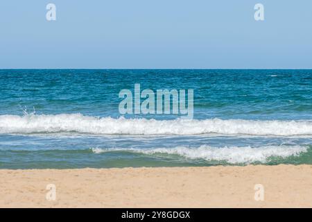 Questa immagine mostra una tranquilla spiaggia con cieli azzurri, un oceano calmo con dolci onde che si infrangono sulla spiaggia sabbiosa. T Foto Stock