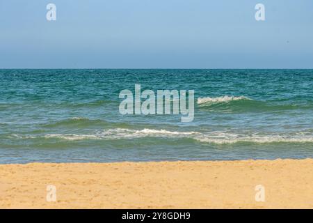 Questa immagine mostra una tranquilla scena di spiaggia con la costa in primo piano, le onde dolci nel mezzo e un cielo azzurro. Foto Stock