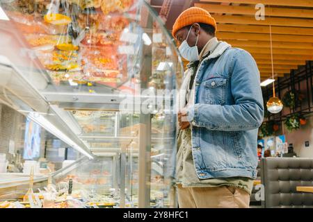 L'uomo in maschera sceglie il cibo dalla finestra e negozi al banco deli durante la pandemia. L'acquirente applica misure di protezione e sicurezza delle malattie in luoghi pubblici Foto Stock