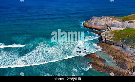 Vista aerea di una costa rocciosa con onde che si infrangono contro la costa. Il vivace oceano blu contrasta con le rocce aspre e l'erba verde sul Foto Stock