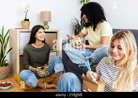 Tre giovani amici diversi mangiano insieme sul divano in un appartamento condiviso Foto Stock