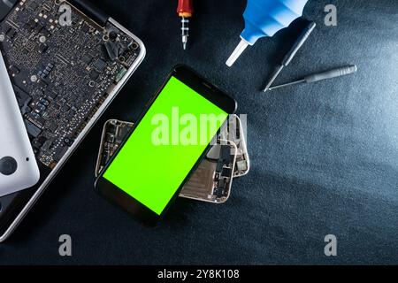 Uno smartphone con uno schermo verde posizionato sopra le parti e gli strumenti smontati del telefono. Lo sfondo è scuro, evidenziando il telefono e gli strumenti utilizzati f Foto Stock