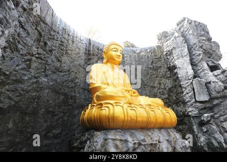 La grande statua dorata del Buddha con cascata e muro di pietra sullo sfondo a Wat Lak si Rat Samoson, Samut Sakhon, Thailandia. Foto Stock