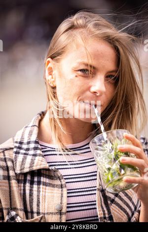 Una giovane donna in una giacca a quadri alla moda si gode un drink all'aperto in una ventosa giornata autunnale, sorseggiando una paglia Foto Stock