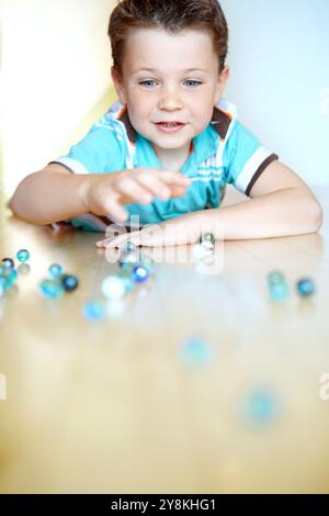 Casa, ragazzo e felice con biglie sorridenti per divertirsi durante la pausa scolastica sul pavimento per attività. Uomo, bambino e concentrazione in appartamento con giocattoli per ga Foto Stock