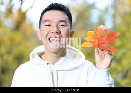 Un giovane uomo sta tenendo in mano una grande foglia d'acero autunnale colorata in un parco forestale all'aperto e sorride alla macchina fotografica Foto Stock