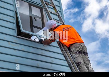 Pittore su una scala che dipinge intorno alla finestra di una vecchia casa in legno/pannelli di legno. Foto Stock