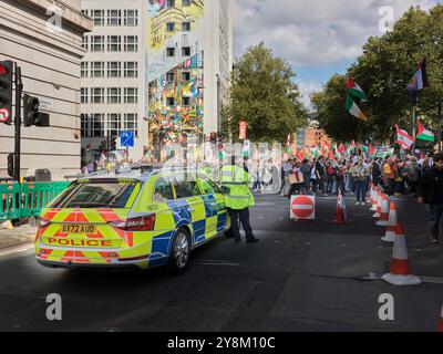 Un ufficiale di polizia sta accanto alla sua auto mentre i manifestanti marciano con le bandiere lungo Southampton Row, Londra, Inghilterra, il 5 ottobre 2024, per sostenere i musulmani di Gaza e Libano nella loro guerra contro gli ebrei in Israele. Foto Stock