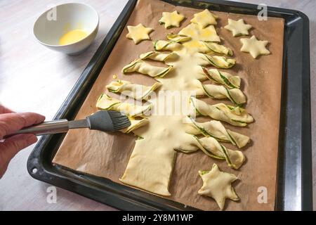 Spalmare il tuorlo sull'impasto di pasta sfoglia ripieno di pesto fatto in casa e tagliarlo a forma di albero di Natale su un vassoio da forno, idea creativa per gli holi stagionali Foto Stock