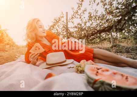 Picnic donna anguria estate - Una donna con un abito rosso si gode un picnic con anguria in un frutteto soleggiato. Foto Stock