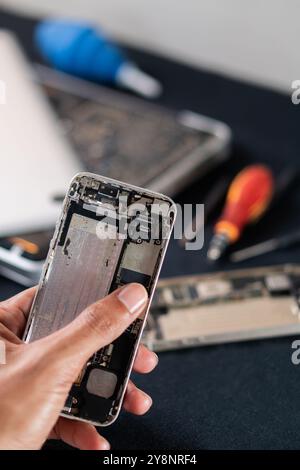 Primo piano di una persona che tiene in mano uno smartphone smontato, rivelando i suoi componenti interni. Lo sfondo presenta strumenti e altri dispositivi elettronici Foto Stock