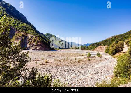 Un ampio e asciutto letto fluviale si estende attraverso una valle nei Monti Pindo, circondata da vegetazione lussureggiante e colline rocciose. Foto Stock