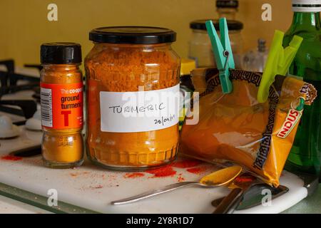 In cucina, la spezia curcuma macinata dai colori vivaci viene decantata in un grande vaso di sottaceti utilizzando un cucchiaino e alcune sono state versate sul piano di lavoro Foto Stock