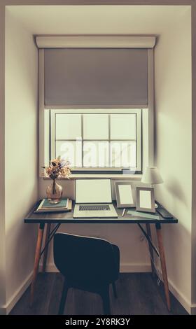 Uno spazio di lavoro minimalista con laptop, libri e cornici fotografiche su una moderna scrivania nera. Un vaso con fiori secchi e una piccola lampada si trovano accanto a una finestra W Foto Stock