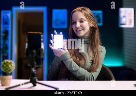 Allegra ragazza di generazione Z in studio domestico che utilizza il telefono per registrare, rivedendo luci mini RGB. Un influencer sorridente filma l'elettronica con lo smartphone su un treppiede, presentando un dispositivo di illuminazione a LED Foto Stock