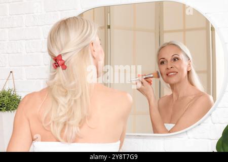 Donna matura che fa il trucco vicino allo specchio in bagno Foto Stock