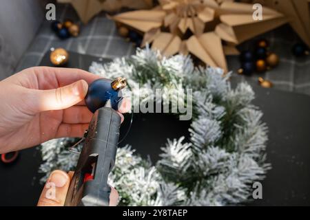 Artigiano con colla calda istruzioni passo dopo passo per realizzare una corona di Natale fatta a mano con decorazioni natalizie nei colori blu e giallo. Mani femminili che creano decorazioni artigianali fatte in casa. Festa di Capodanno. Vacanze invernali fase 2 Foto Stock